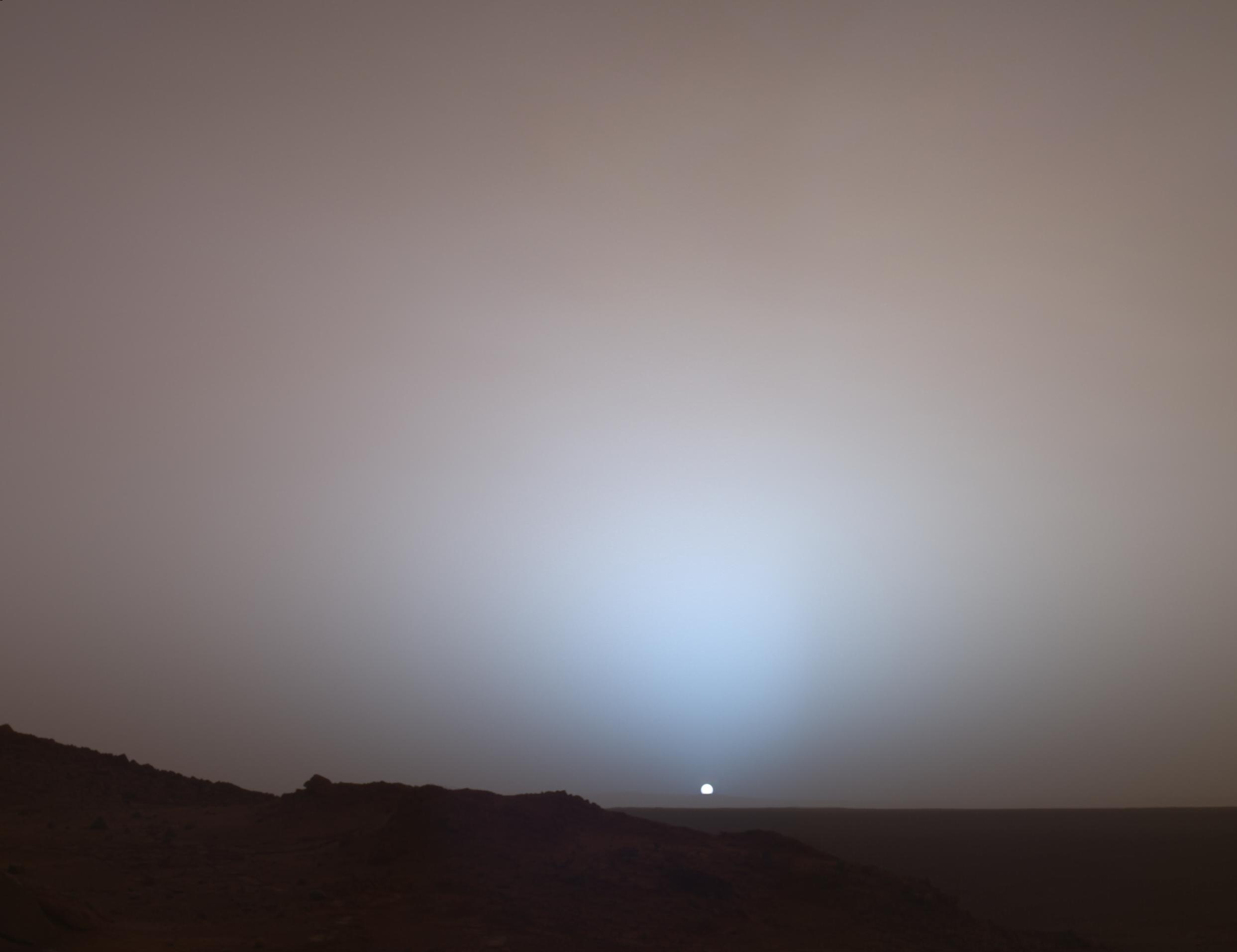 A small Sun sets behind the dark rim of Gusev crater beneath a blue glow in the Martian sky.